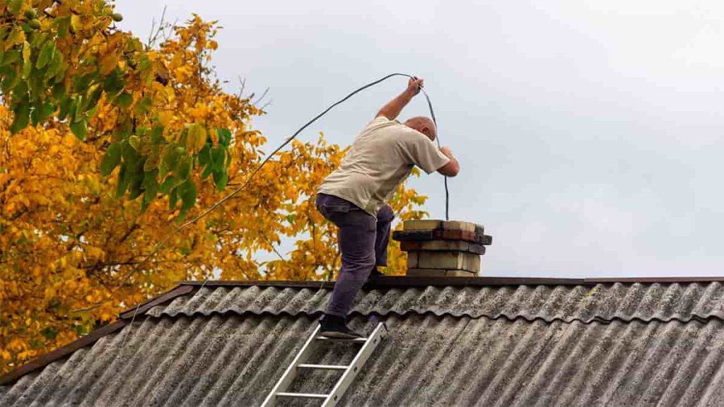 Een vakman die op een ladder op een dak staat en een schoorsteen veegt met een lange borstel.