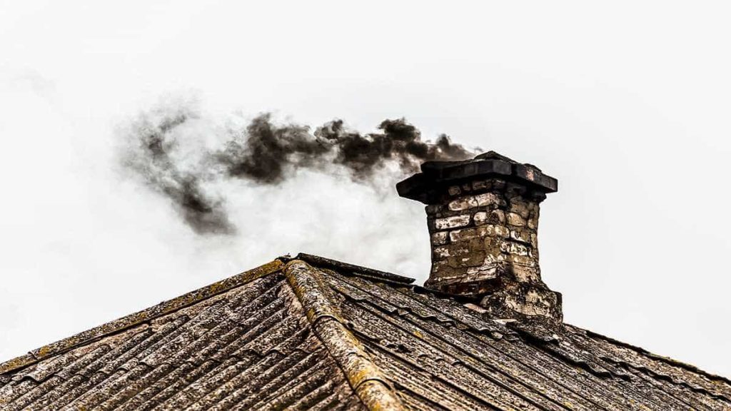 Een schoorsteen op het dak van een oud huis, met droog mos aan de buitenkant. Er komt zwarte rook uit de schoorsteen.